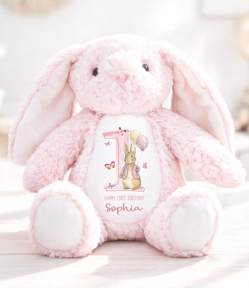 May include: A pale pink plush bunny toy with a white belly. The bunny features a graphic of a rabbit holding balloons, the number 1, butterflies, and the text "Happy First Birthday Sophia".