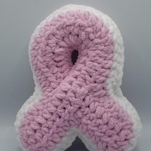 May include: A pink and white crocheted ribbon pillow. The pillow is shaped like a ribbon and is made with a soft, fluffy yarn.