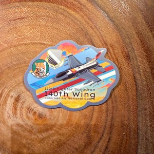 May include: A colorful sticker featuring a fighter jet flying through a cloudy sky. The sticker has the text "120th Fighter Squadron 140th Wing Colorado Air National Guard".