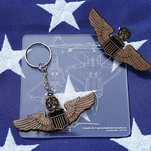 May include: Wooden pilot wings keychain and pin. The wings are brown with black accents, and the keychain has a silver ring. The background is a blue fabric with white stars, resembling the American flag. A clear plastic card with an airplane blueprint is also present.