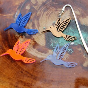 May include: Four hummingbird bookmarks, one silver, one brown wood, one blue, and one orange. The bookmarks are on a wooden surface.