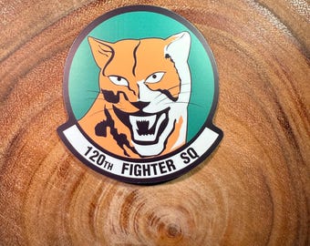 May include: A green, white, and orange round sticker with a snarling tiger face and the text "120th Fighter SQ".