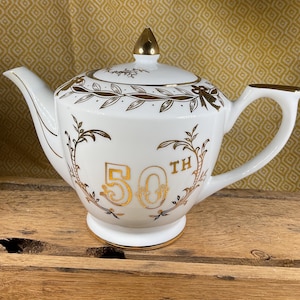 May include: White ceramic teapot with gold accents, including the number "50" and the word "TH" in gold script, celebrating a 50th anniversary. The teapot has a gold handle and a gold finial on the lid.