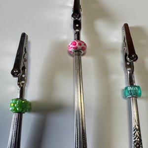 May include: Three silver-toned alligator clip holders, each adorned with a decorative bead. One has a green bead, another a pink and white spotted bead, and the third a teal bead. The holders are made from repurposed silverware.