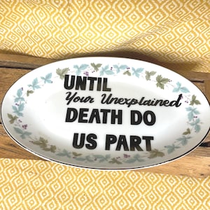May include: Oval ceramic dish with the text "UNTIL Your Unexplained DEATH DO US PART" in black. The dish has a white background with a black rim and is decorated with green and blue floral accents.