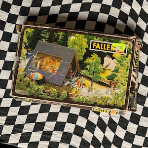 Puede incluir: Una caja de kit de modelo Faller vintage con una escena de cabaña en forma de A en miniatura con árboles, figuras y un paraguas colorido. La caja está desgastada, con el logotipo de Faller visible. La escena incluye un pequeño puente y una bicicleta.