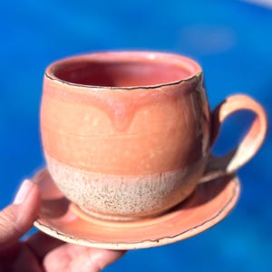 May include: A peach-coloured ceramic teacup and saucer set with a speckled glaze. The cup has a rounded shape and a handle. The saucer has a scalloped edge.