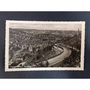 May include: A black and white postcard showing an aerial view of a city with a road winding through it. The text on the postcard reads "Stuttgart Neue Weinsteige 247".