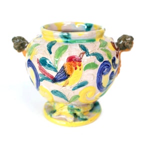 May include: A yellow ceramic vase with a bird and floral design. The vase has two handles shaped like animal heads.