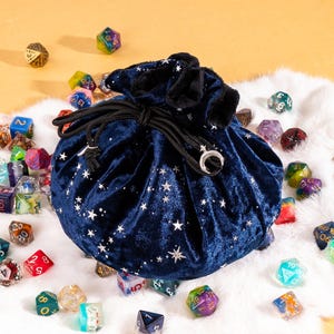 May include: A dark blue velvet drawstring pouch featuring a silver star pattern and a crescent moon charm on the cord ends, surrounded by a collection of multi-coloured polyhedral dice scattered across a white faux fur surface.