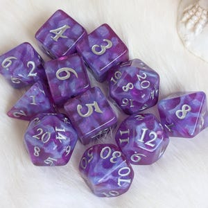 Peut inclure: Un ensemble de dés polyédriques violets et blancs marbrés avec des chiffres argentés. L'ensemble comprend des dés de différentes formes, comme des dés à six, huit, dix, douze et vingt faces, utilisés dans les jeux de rôle.