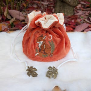 May include: An orange drawstring pouch with a fox embroidery design. The pouch has a cream-colored interior and white drawstrings with leaf-shaped metal accents. The fox is embroidered in brown and white, with a crescent moon and foliage.