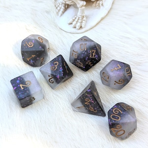May include: Set of seven polyhedral dice in shades of gray, black, and purple with gold numbering. The dice are made of a translucent material with glitter and are designed for tabletop role-playing games.