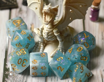 May include: A set of blue and gold dice with glitter and paw prints. The dice are scattered around a white skeletal dragon figurine.