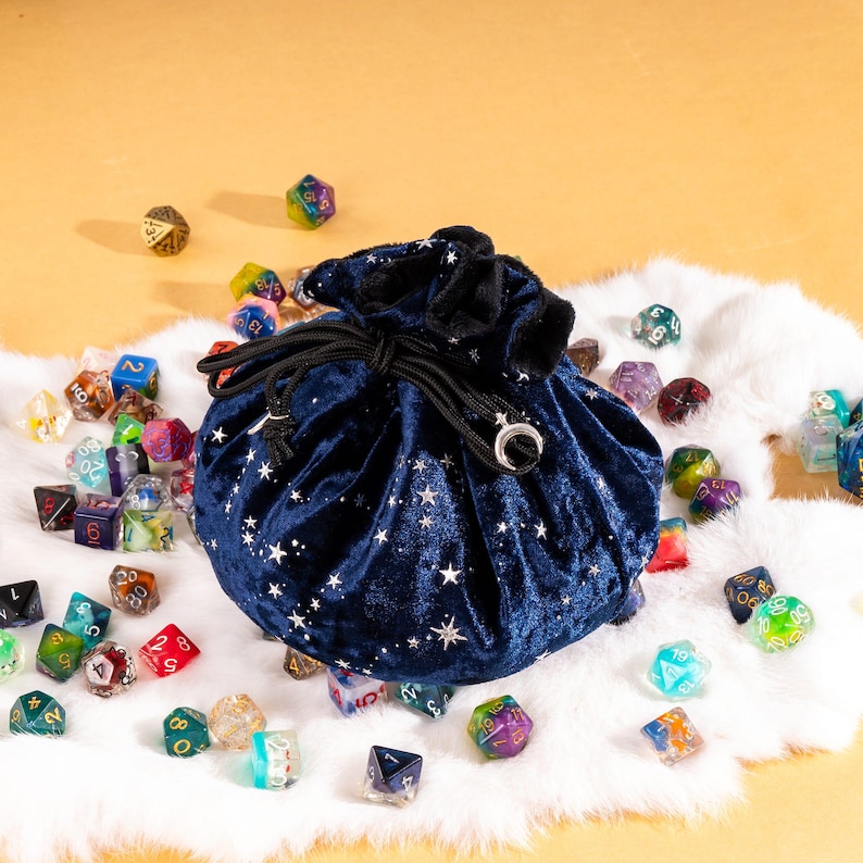 May include: A dark blue velvet drawstring pouch featuring a silver star pattern and a crescent moon charm on the cord ends, surrounded by a collection of multi-colored polyhedral dice scattered across a white faux fur surface.