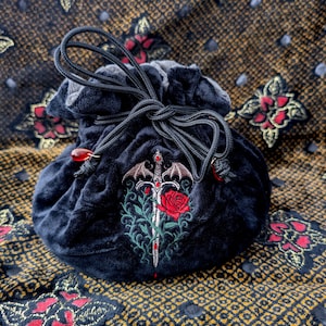 May include: A dark blue velvet drawstring bag with an embroidered design featuring a sword, rose, and bat wings. The bag has a black cord drawstring and red bead accents. The background is a patterned fabric with red and gold floral designs.