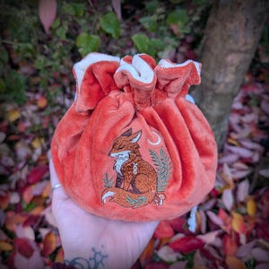 May include: A plush orange drawstring bag with a white interior. The bag features an embroidered design of a fox, a crescent moon, and foliage. The bag is held in a hand against a blurred background of fall leaves.
