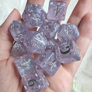 May include: A set of ten purple and white dice with silver glitter and numbers. The dice have a variety of symbols including a crescent moon, a star, and a cross.