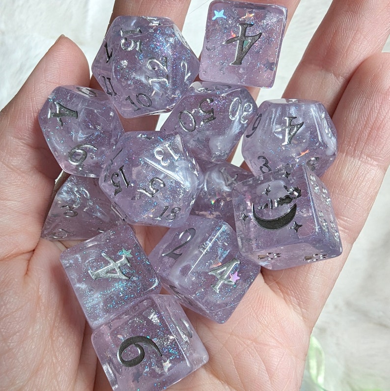 May include: A set of ten purple and white dice with silver glitter and numbers. The dice have a variety of symbols including a crescent moon, a star, and a cross.