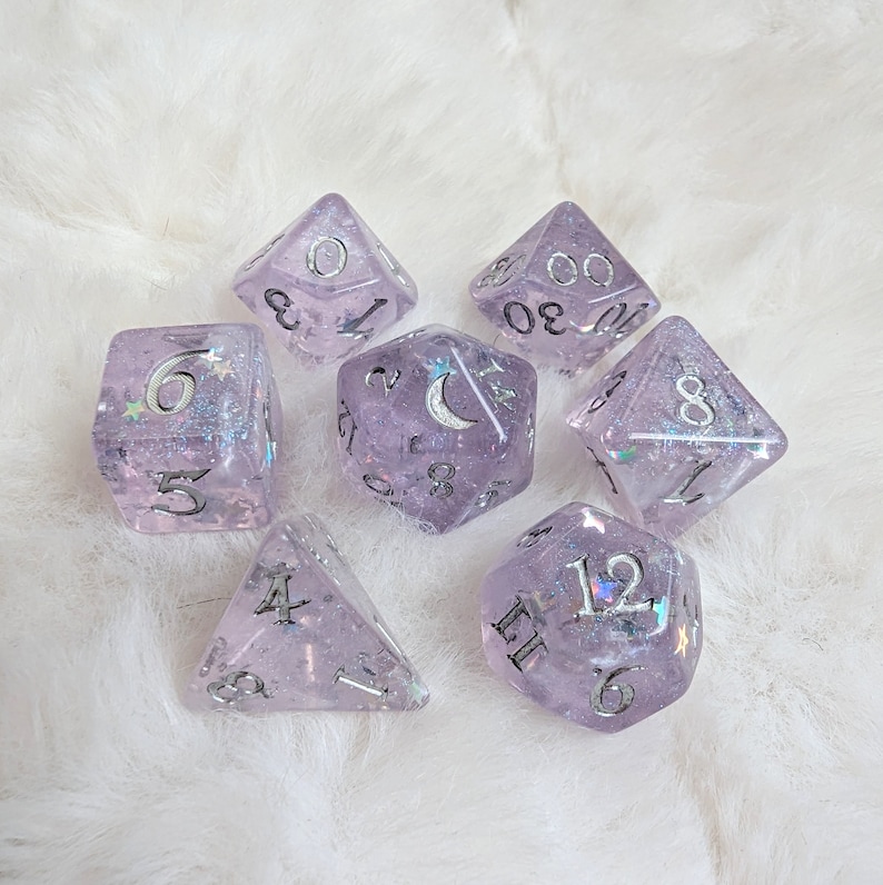 May include: Set of seven iridescent purple dice with silver glitter and white numbers. The dice are shaped like a d4, d6, d8, d10, d%, d12, and d20.