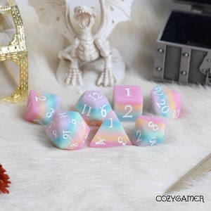 May include: A set of seven pastel-colored dice with white numbers. The dice are made of a translucent material and have a rainbow swirl pattern. The dice are arranged in a circle on a white fluffy surface.