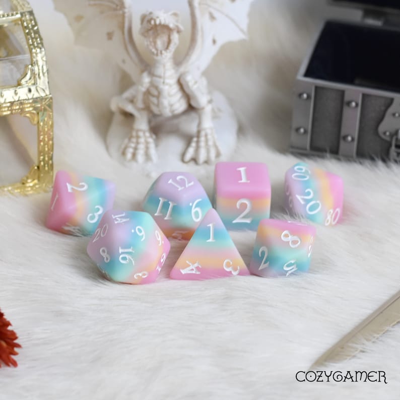 May include: A set of seven pastel-colored dice with white numbers. The dice are made of a translucent material and have a rainbow swirl pattern. The dice are arranged in a circle on a white fluffy surface.