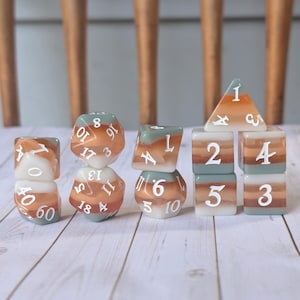 Puede incluir: Un conjunto de dados poliédricos de varias formas, incluyendo un d20, d12, d6 y d4. Los dados presentan un diseño en capas con colores blanco, naranja y turquesa. Los números son blancos.