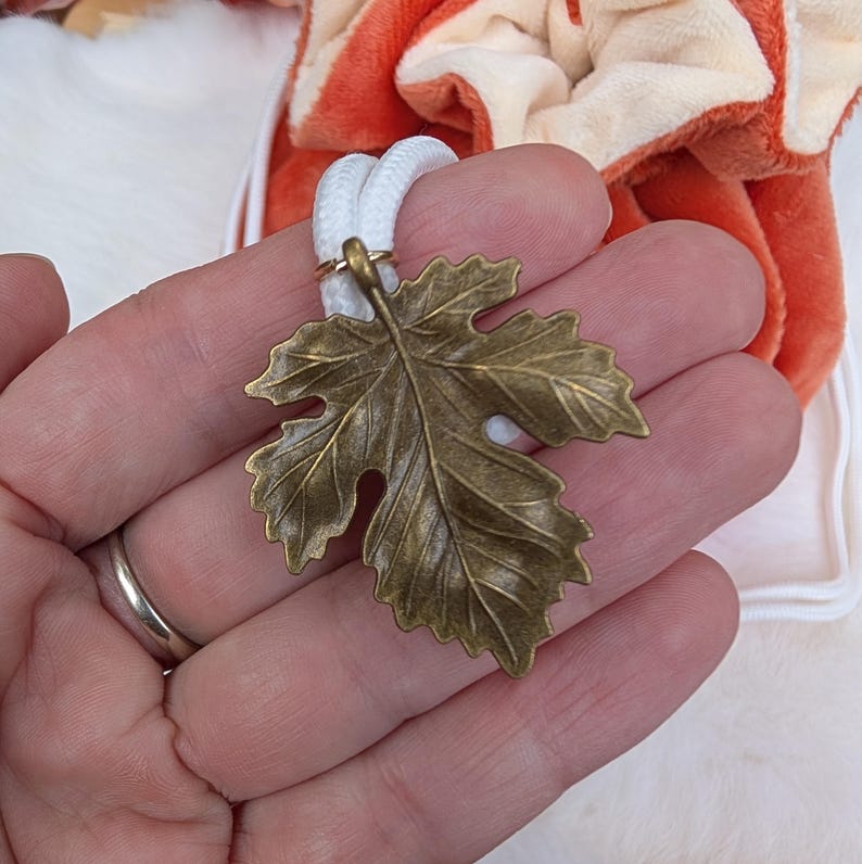May include: A bronze-colored leaf pendant with a white cord. The leaf has detailed veining and is attached to the cord with a small gold-colored ring. The pendant is held in a hand.