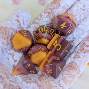 May include: A set of eight-sided dice with a purple and orange swirl pattern. The dice have gold numbers on each side. The dice are on a white lace surface.