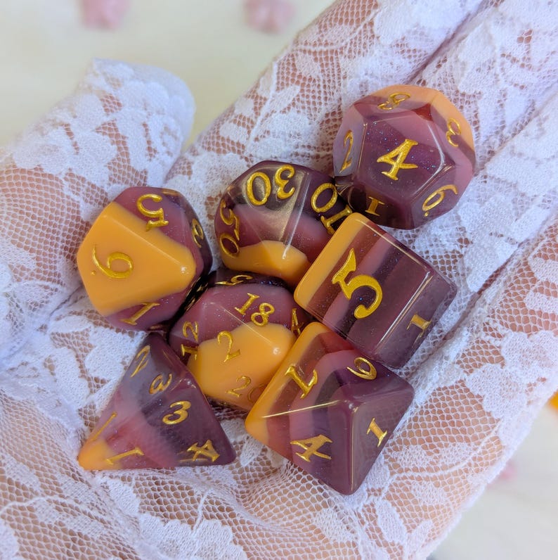 May include: A set of eight-sided dice with a purple and orange swirl pattern. The dice have gold numbers on each side. The dice are on a white lace surface.