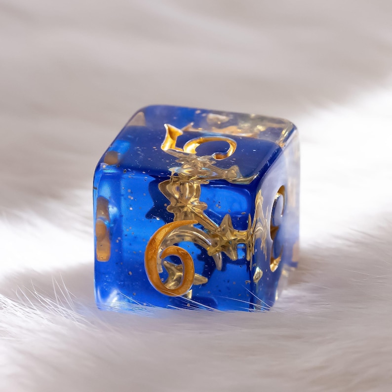 May include: A six-sided die with a transparent blue resin body. The die features gold-colored ornate designs and numerals. The number 6 is visible on the face. The die is designed for tabletop games.