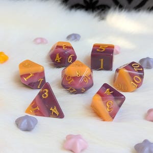 May include: Set of ten-sided dice with a gradient of purple and orange. The dice have gold numbers.