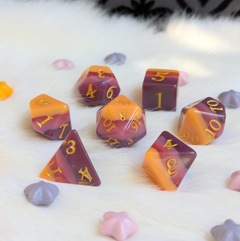 May include: Set of ten-sided dice with a gradient of purple and orange. The dice have gold numbers.