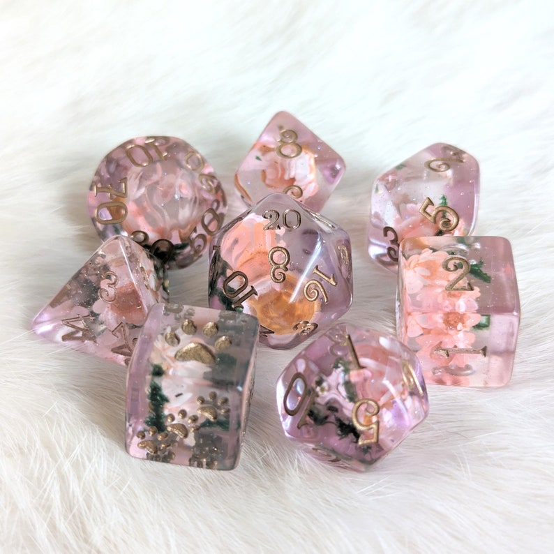 May include: A set of seven pink and gold dice with floral and other nature-inspired designs. The dice are made of clear resin with embedded flowers and other natural elements.