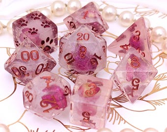 May include: A set of seven polyhedral dice made of clear resin containing pink floral inclusions and fine glitter. The dice feature metallic copper-colored numbers and paw print symbols on the six-sided die, arranged in a cluster for tabletop gaming.