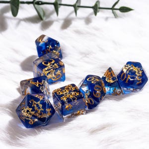 May include: Set of eight polyhedral dice in various shapes. The dice are a deep blue with gold-colored numbers and decorative elements. The dice are arranged on a white, fluffy surface.