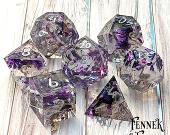 May include: Set of seven clear resin dice with purple and white flower accents. The dice are numbered 1-6, 1-20, and 4-sided. The dice are on a light wood background with the text "Fennel & Finch Dice Co."