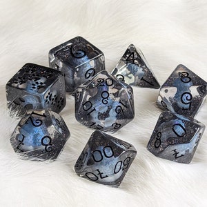 May include: A set of seven polyhedral dice. Each die is clear with a dark blue and black swirling pattern and black numbering. The dice are arranged on a white, fluffy surface.