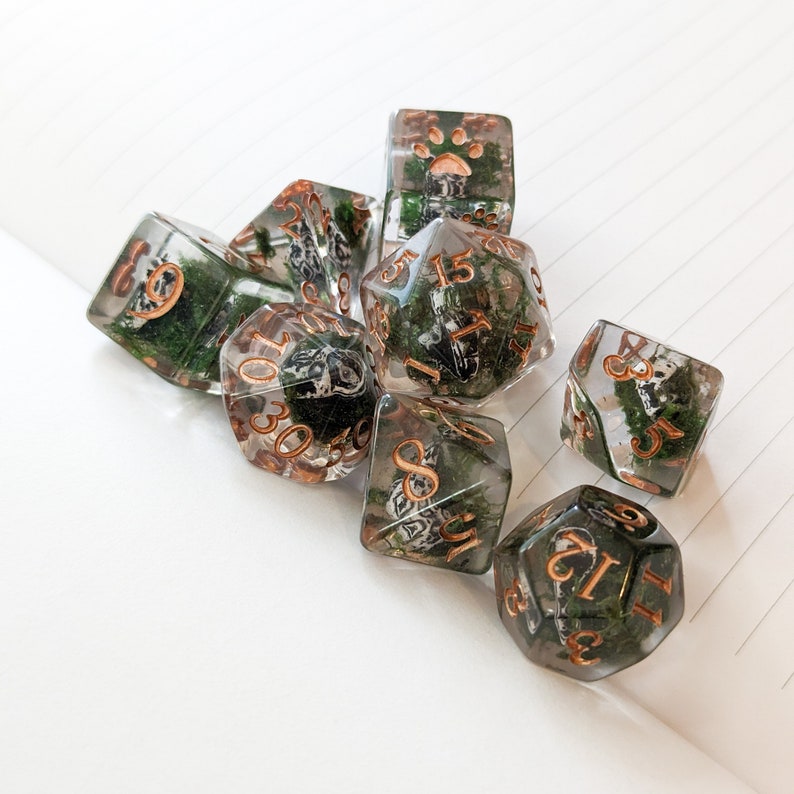 May include: A set of clear resin dice with embedded green moss and copper-colored numbers. The dice are in various shapes, including a d20, d12, and d6, perfect for tabletop gaming.