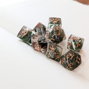 May include: A set of clear resin dice with green moss inclusions and copper-colored numbering. The dice are in various polyhedral shapes, including D6, D8, and D20, and are arranged on a white surface. The dice are designed for tabletop gaming.