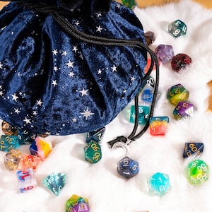 May include: A dark blue velvet drawstring bag featuring a silver star pattern, accompanied by a collection of multi-colored polyhedral dice. The bag includes a silver crescent moon charm attached to the black cord drawstring closure.
