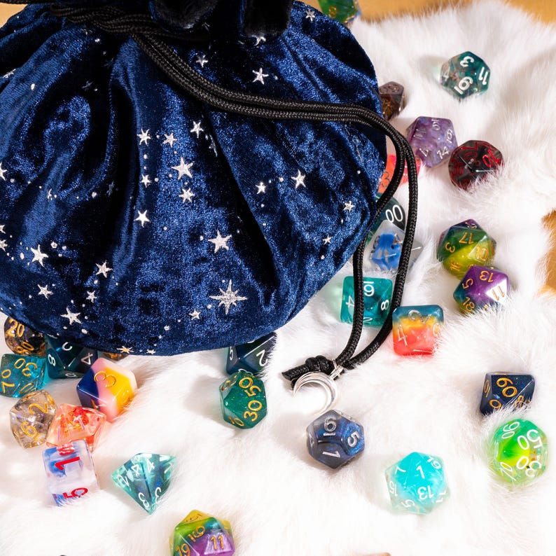 May include: A dark blue velvet drawstring bag featuring a silver star pattern, accompanied by a collection of multi-colored polyhedral dice. The bag includes a silver crescent moon charm attached to the black cord drawstring closure.
