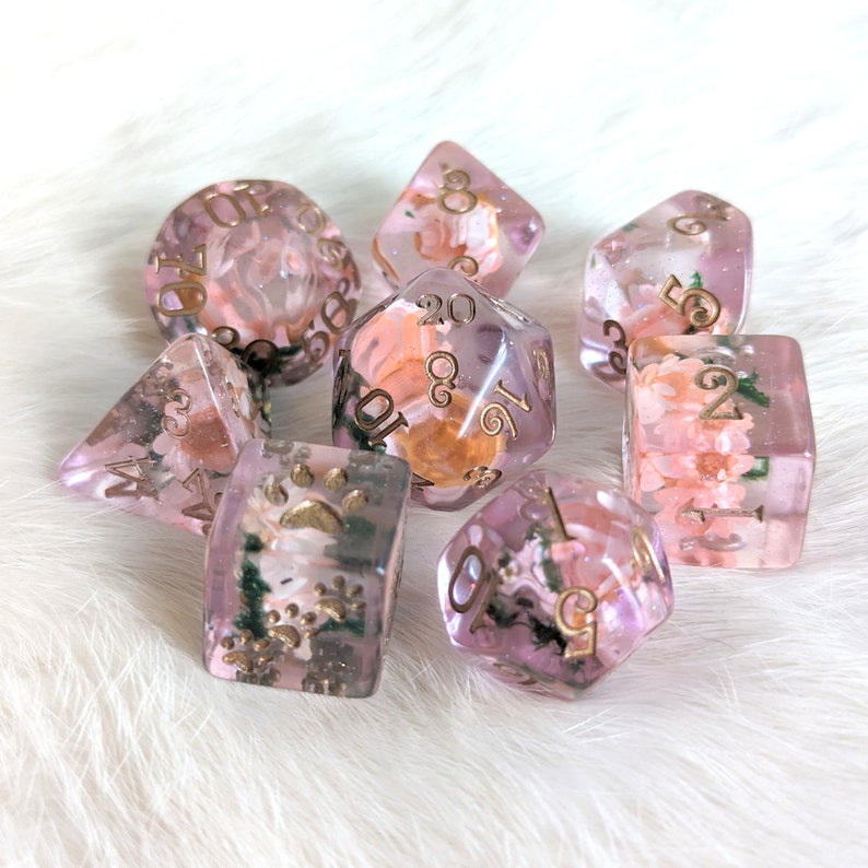 May include: A set of seven pink and gold dice with floral and animal designs. The dice are translucent and have gold numbers on them.