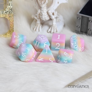 May include: A set of seven pastel-colored dice with white numbers. The dice are a mix of standard polyhedral shapes, including a d4, d6, d8, d10, d12, and d20. The dice are on a white fluffy surface.