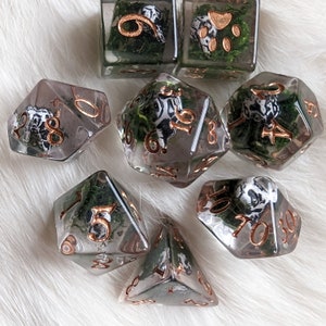 May include: A set of clear resin dice with copper-colored numbering. Each die features a black and white design and is filled with green moss. The dice are in various geometric shapes, including a cube, a d20, and a d10.