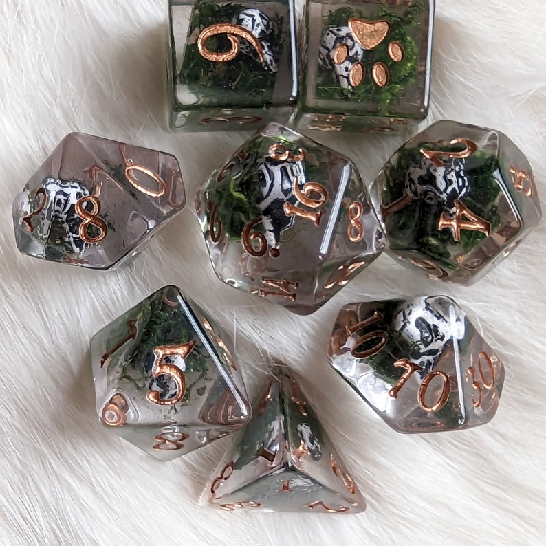 May include: A set of clear resin dice with copper-colored numbering. Each die features a black and white design and is filled with green moss. The dice are in various geometric shapes, including a cube, a d20, and a d10.
