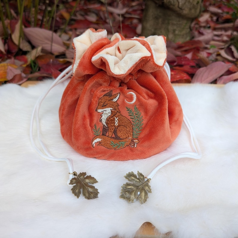 May include: An orange drawstring pouch with a fox embroidery design. The pouch has a cream-colored interior and white drawstrings with leaf-shaped metal accents. The fox is embroidered in brown and white, with a crescent moon and foliage.