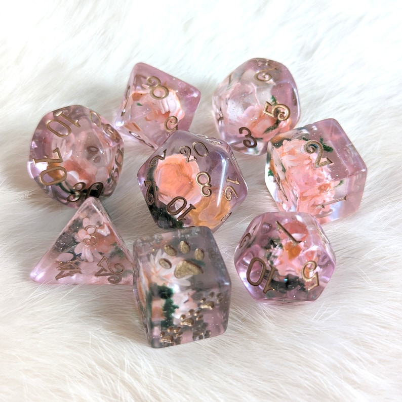 May include: A set of eight pink and gold dice with floral and paw print designs. The dice are translucent and have gold numbers.