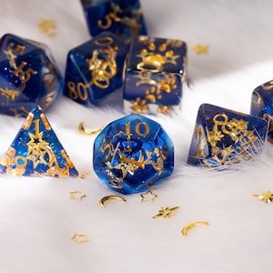 May include: A collection of blue and gold resin dice with gold numbering and decorative accents. The dice are in various shapes, including a pyramid, cube, and a 20-sided die. Small gold stars and crescent moon shapes are scattered around the dice.
