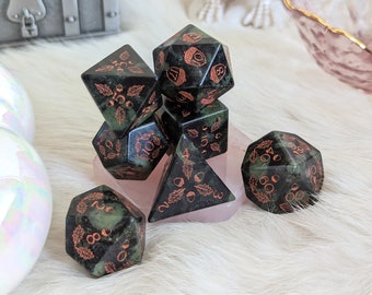 May include: Set of seven green and black dice with copper acorn designs. The dice are on a pink and white stone base.